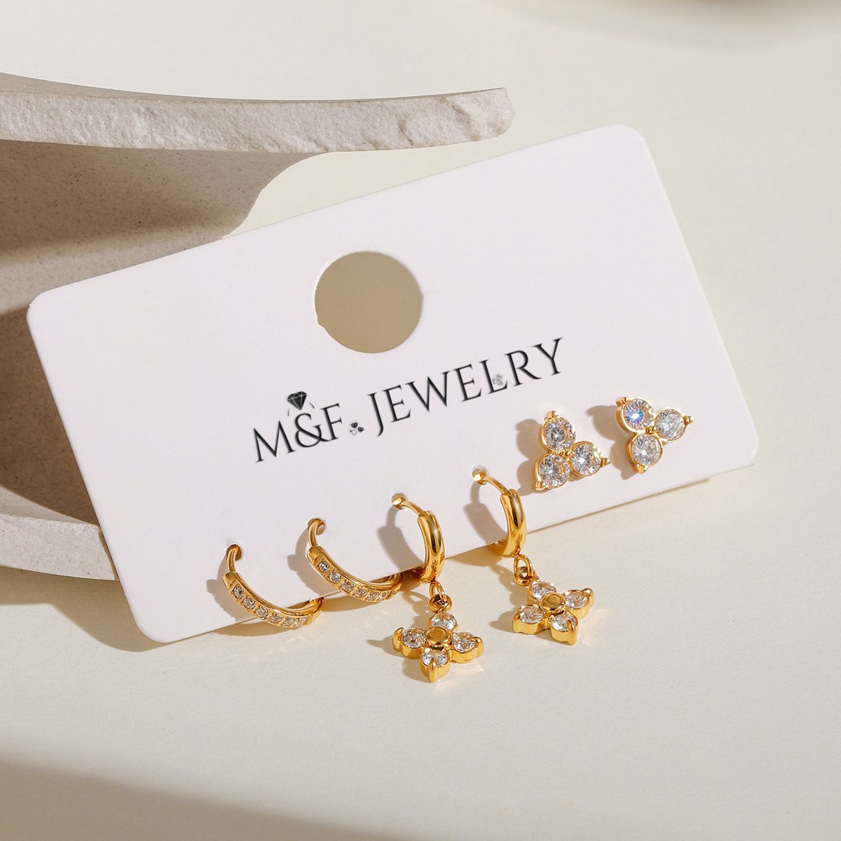 Three styles of gold earrings—floral dangle, triple stone studs, and CZ hoops—arranged in natural light