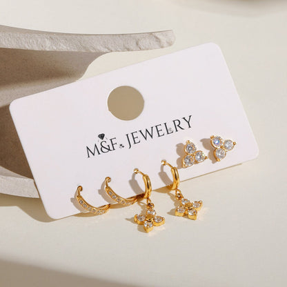 Three styles of gold earrings—floral dangle, triple stone studs, and CZ hoops—arranged in natural light