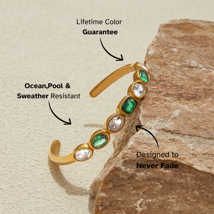 Gold cuff bracelet with emerald and clear stones, styled with natural stone props.