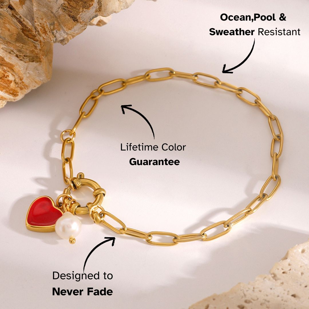 Golden link bracelet with red heart and pearl charms, resting on marble stone