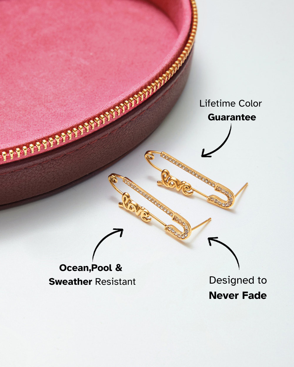 Two gold safety pin earrings with "Love" spelled out in crystals, resting in a pink jewelry box.