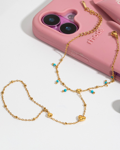 Gold chain with heart and turquoise bead details in front of a pink phone case.