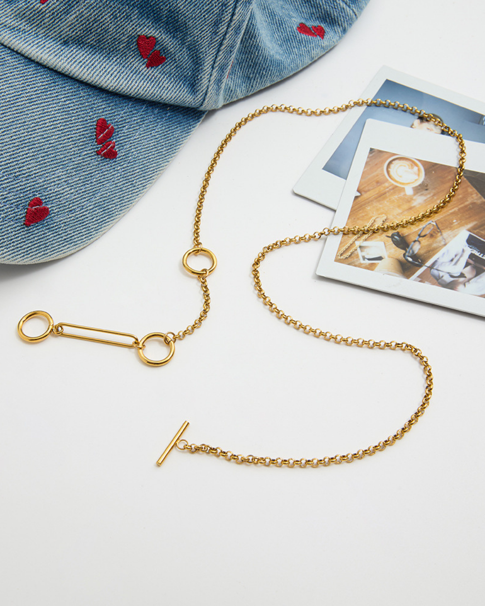 Gold chain necklace with safety pin clasp on a white surface with denim cap and photos.