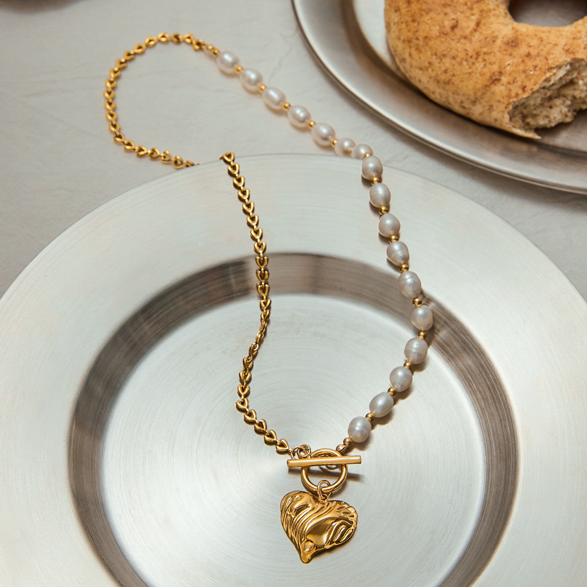 Stylish asymmetric necklace with baroque pearls and hammered gold heart charm resting on a silver-toned plate