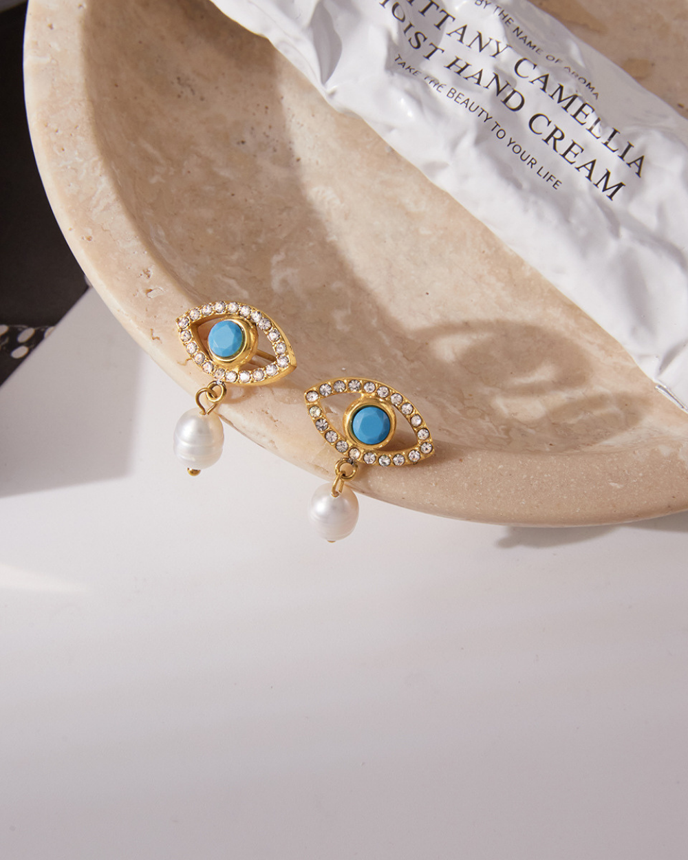 Elegant gold evil eye earrings with blue center and pearl charm displayed on marble jewelry dish.