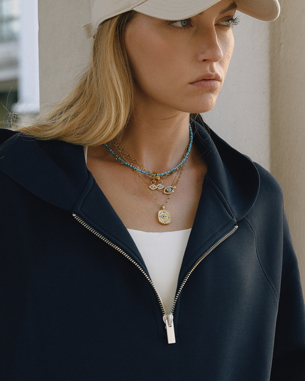 Woman wearing a navy jacket and beige cap with layered necklaces.