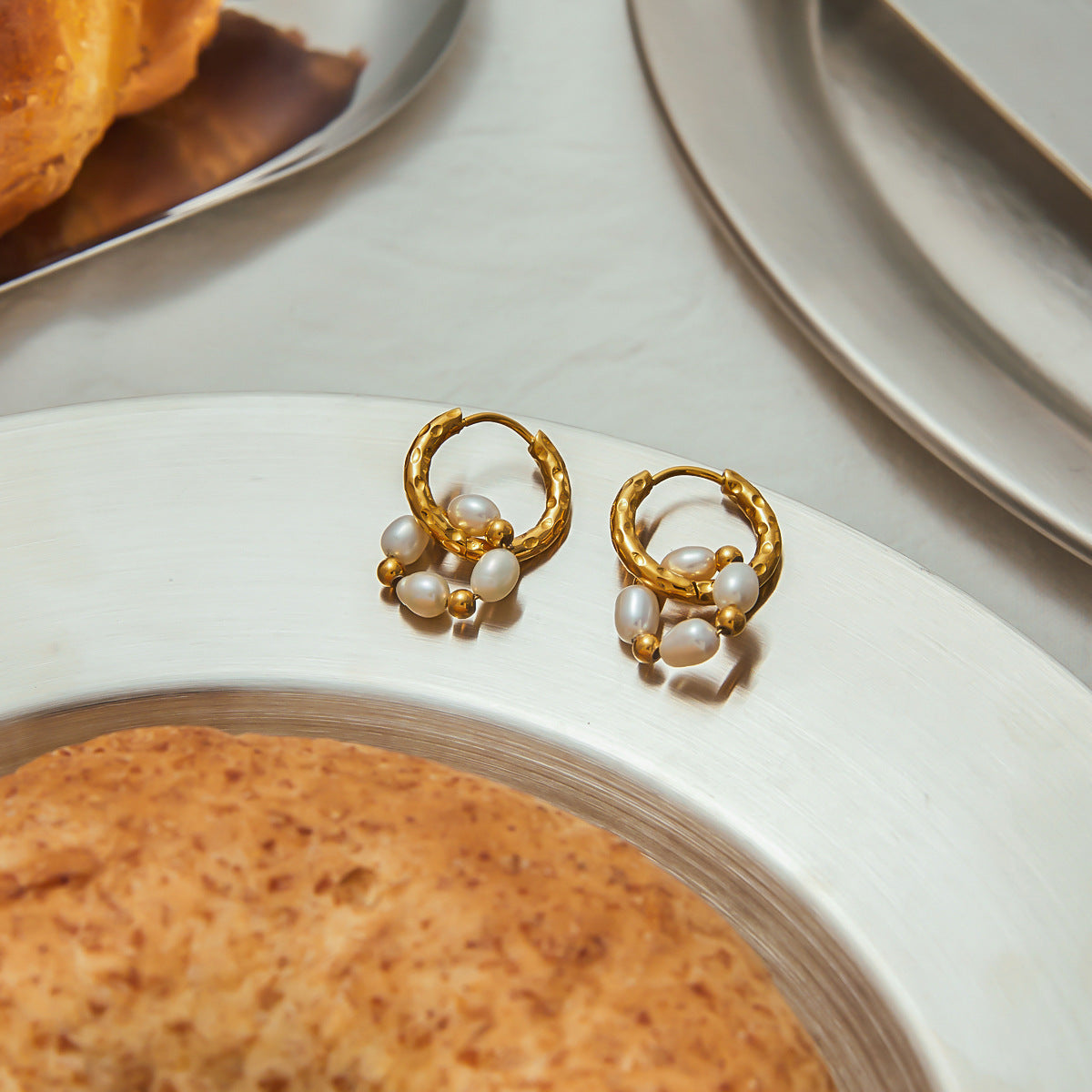 Golden hoop earrings with pearl charms placed on a polished table setting next to bread, evoking relaxed elegance.