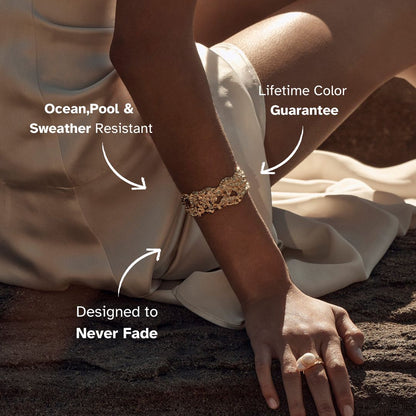 Model wearing a sculpted gold bracelet while reclining on sand at sunset.