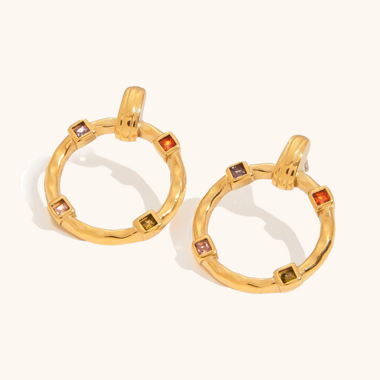 Round gold earrings with multi-colored gemstones laid on stone surface