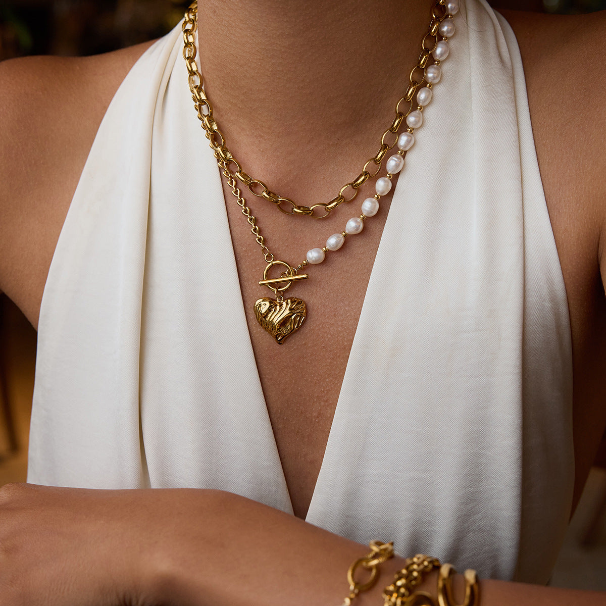 Model wearing heart charm necklace with asymmetric pearl and chain design, styled with gold bracelets