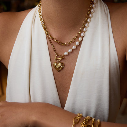 Model wearing heart charm necklace with asymmetric pearl and chain design, styled with gold bracelets