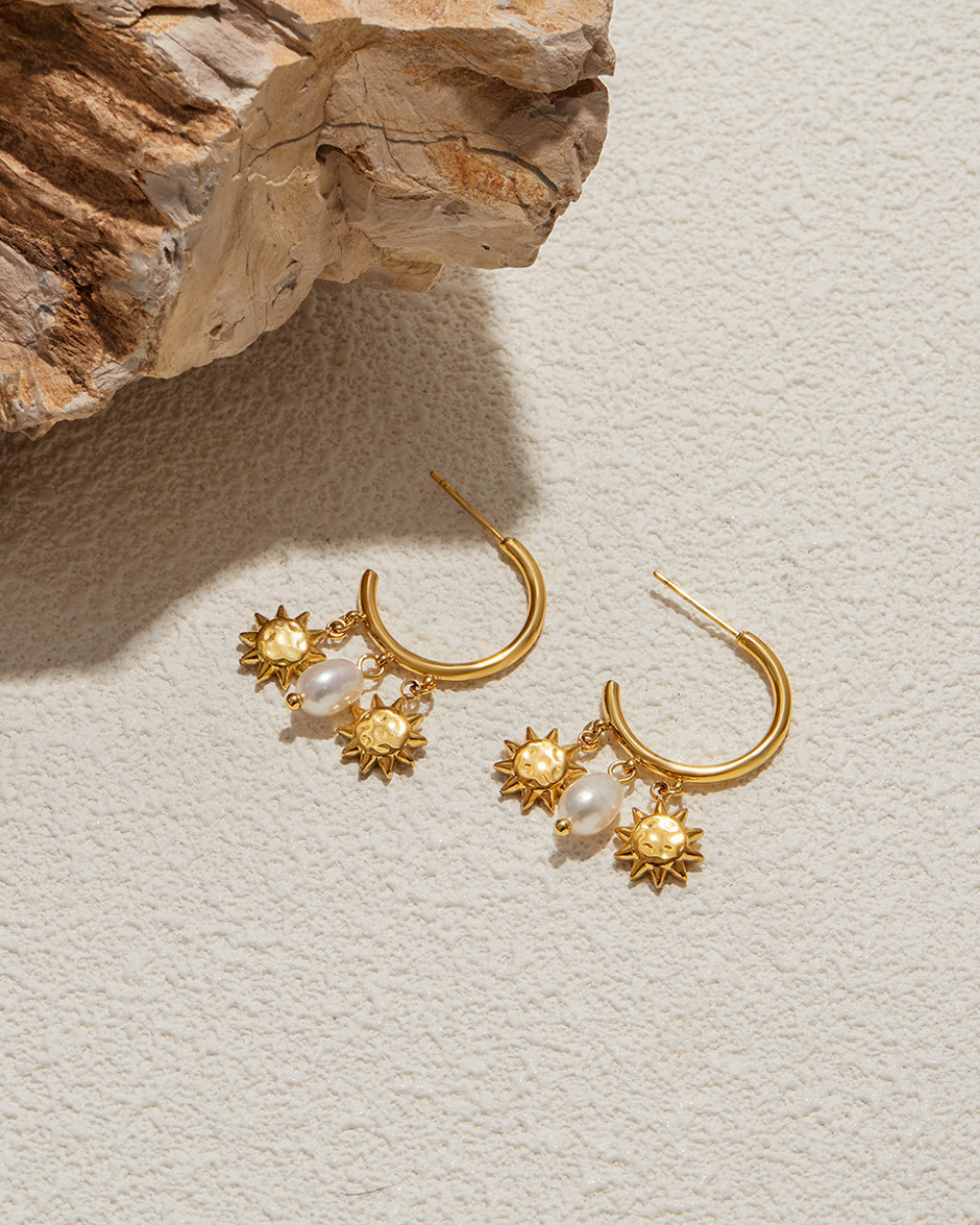 Gold earrings with sun and pearl designs on a textured surface