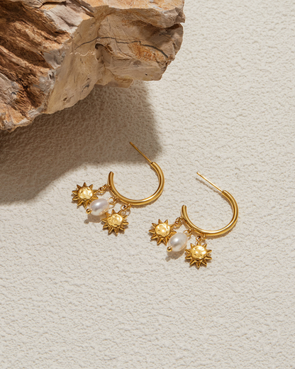Gold earrings with sun and pearl designs on a textured surface