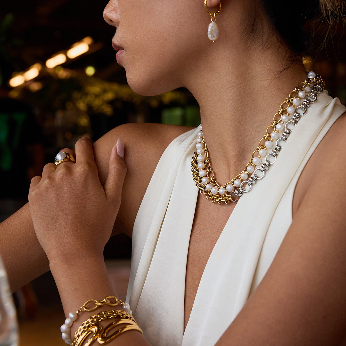 Woman wearing gold and silver chunky chain necklace with pearl strands, styled with matching pearl earrings at a dinner scene.