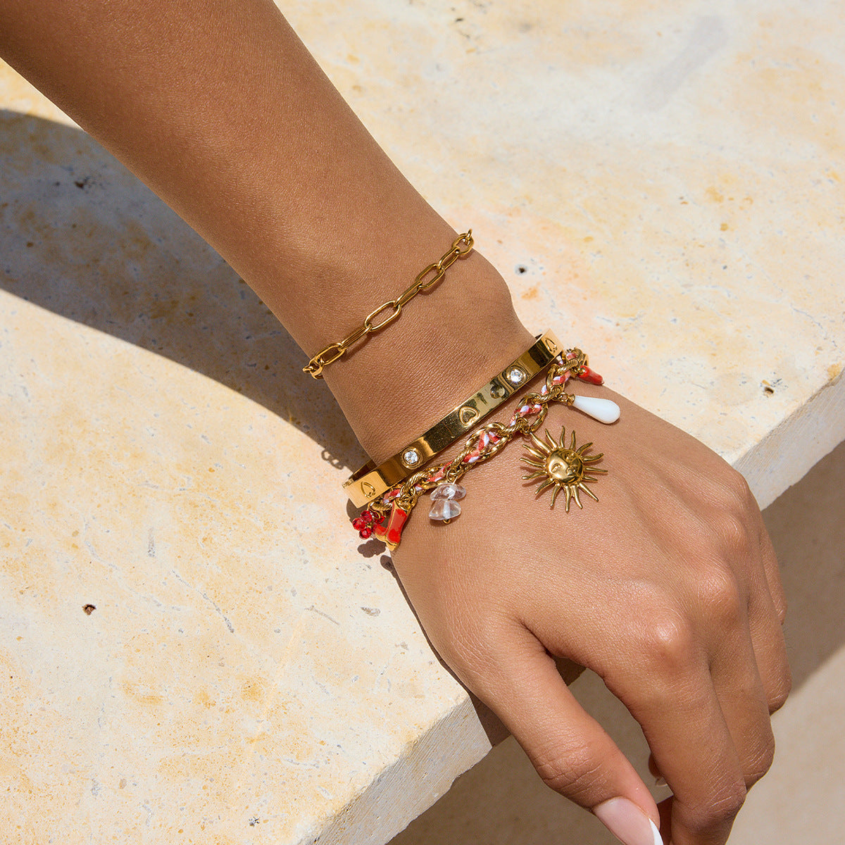 Hand wearing multiple gold bracelets with charms on a light stone surface