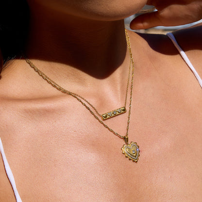 Model wearing layered gold necklaces with heart and bar pendants on sunlit skin