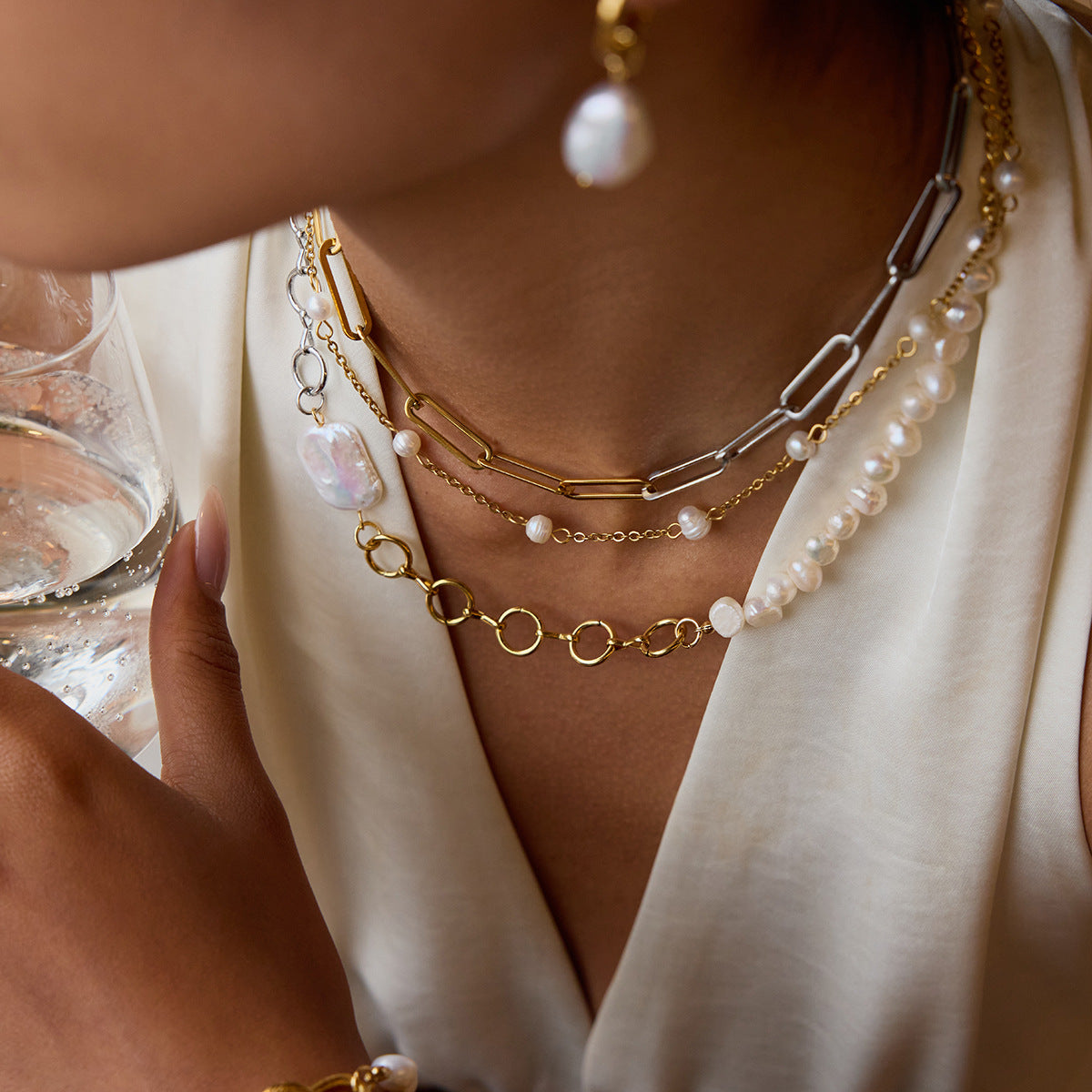 Woman layering asymmetrical pearl necklace with other dainty chains and holding a glass
