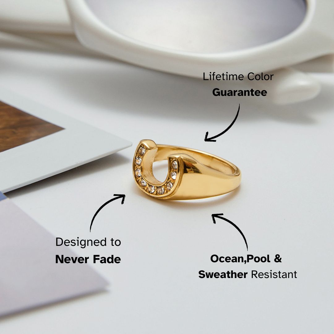 Gold horseshoe ring with crystals shown on a white tabletop beside sunglasses and photos