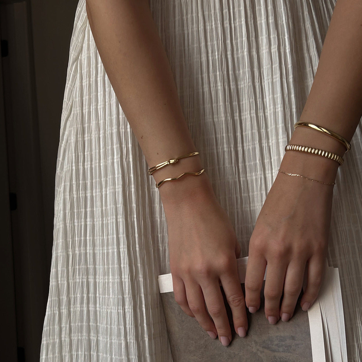 Model’s hand wearing gold beaded bracelet stacked with solid bangle, lifestyle scene with book