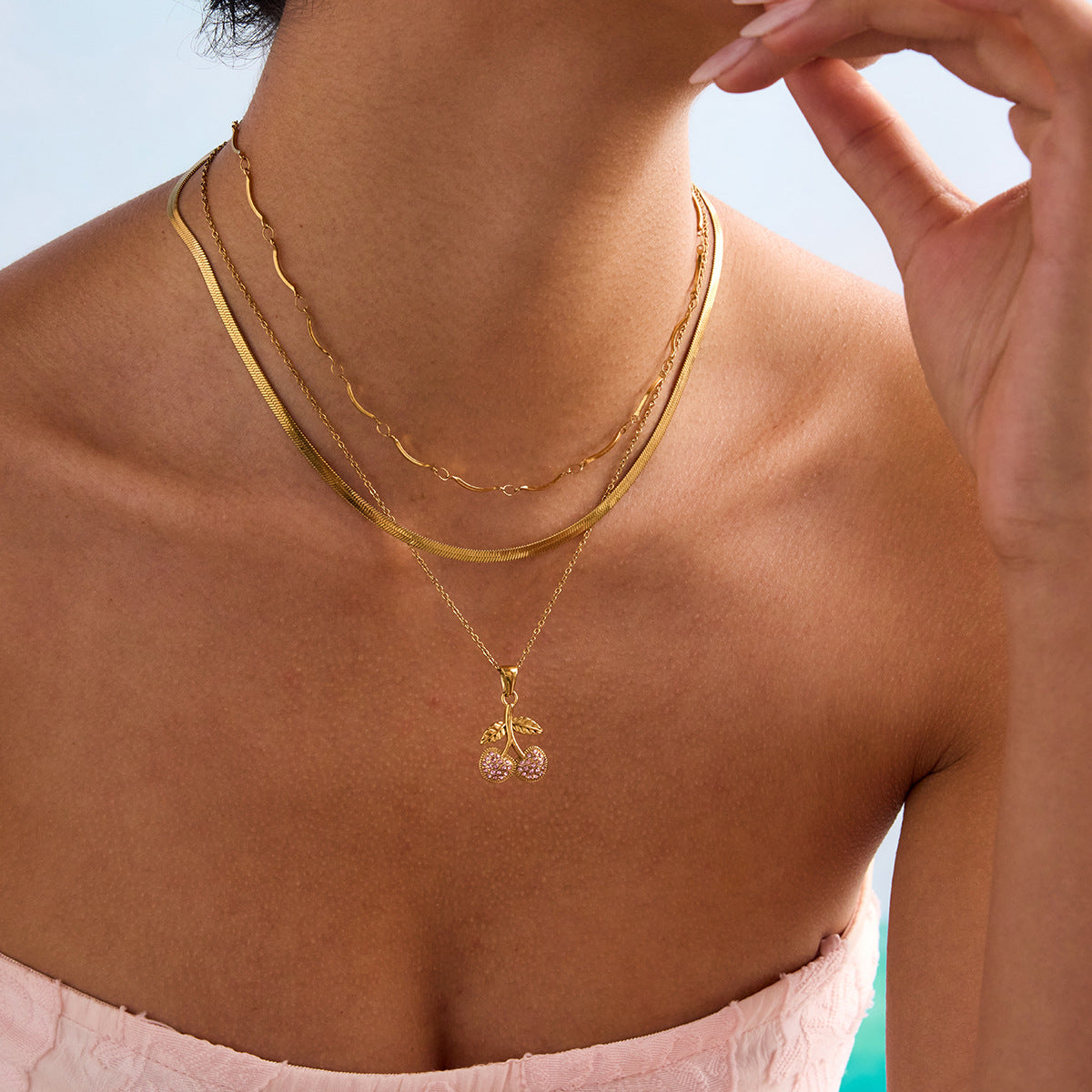 Close-up of layered gold necklaces styled with cherry pendant and ring, elegant summer look.