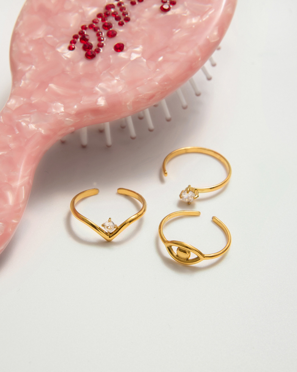 Flat lay of three gold stacking rings including evil eye design and zircon rings placed beside a pink pearl comb.