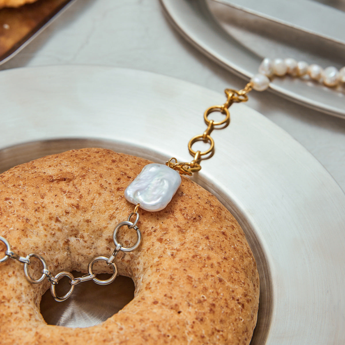 Close-up of two-tone chain pearl necklace styled with a rustic bagel on a silver plate