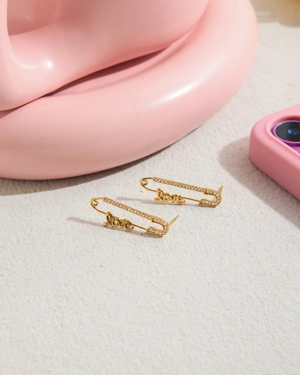 Two gold "Love" safety pin earrings on a textured white surface next to a pink decorative object, under bright light.