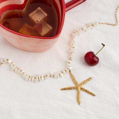 Playful summer necklace next to heart-shaped cup and cherry on linen background