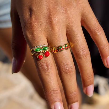 Colorful fruit-inspired gold rings shown on a model's hand in sunlight