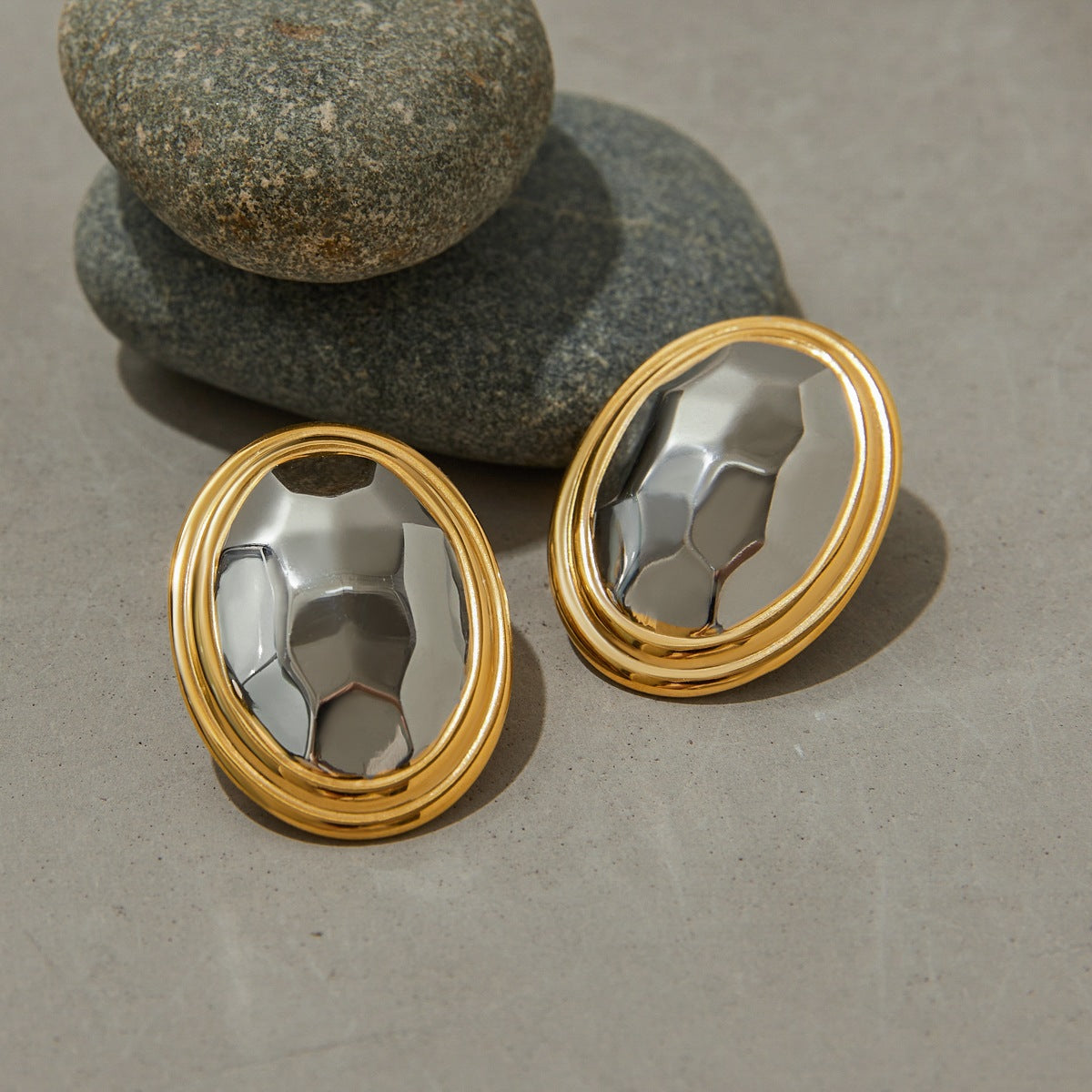 Pair of gold-rimmed silver oval earrings resting on smooth stones, showcasing reflective hammered texture