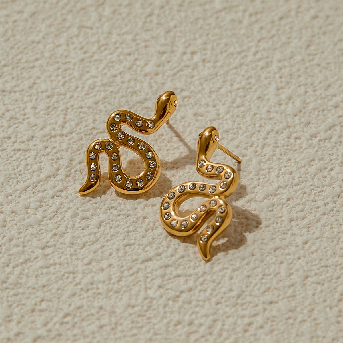 Gold snake-shaped earrings with pavé crystals styled on sandy beige surface