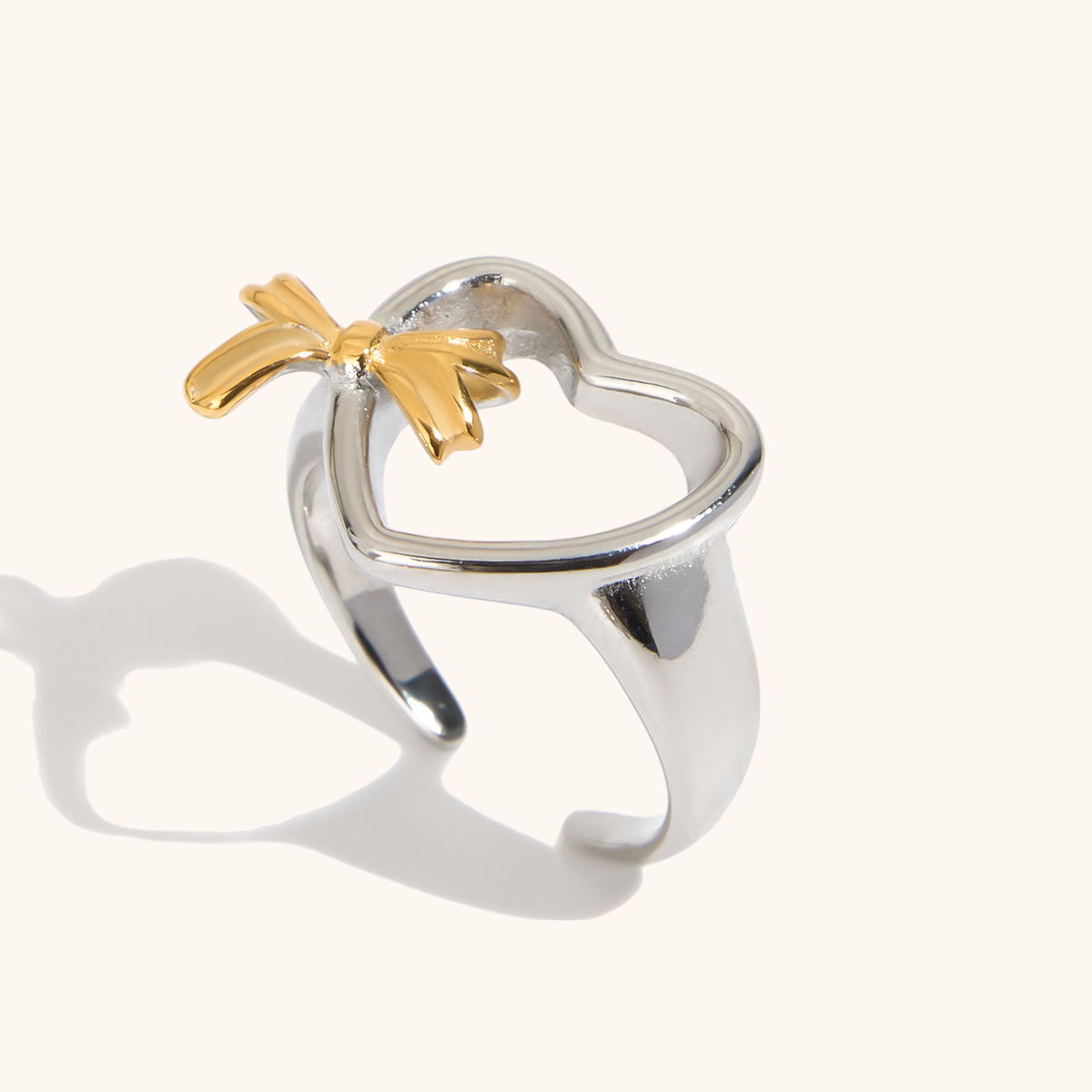 Close-up of silver heart-shaped open ring with gold bow charm, styled on white background.