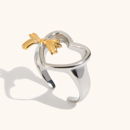 Close-up of silver heart-shaped open ring with gold bow charm, styled on white background.
