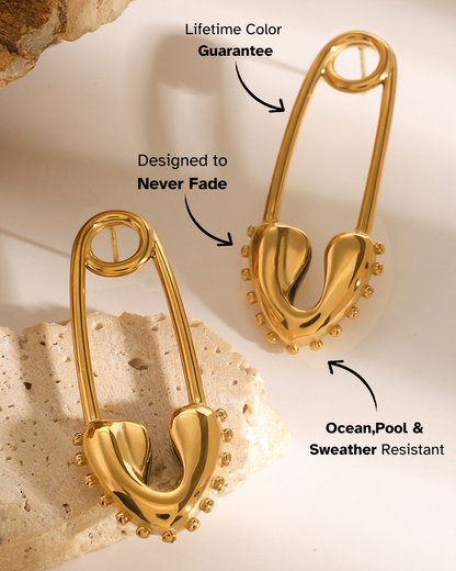 Gold safety pin earrings on a textured beige background with text about color guarantee and resistance.