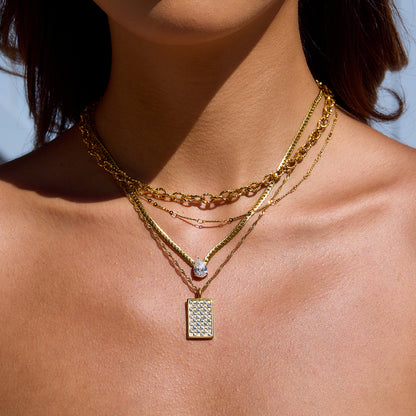 Model wearing layered gold necklaces featuring a square grid pendant in sunlight.