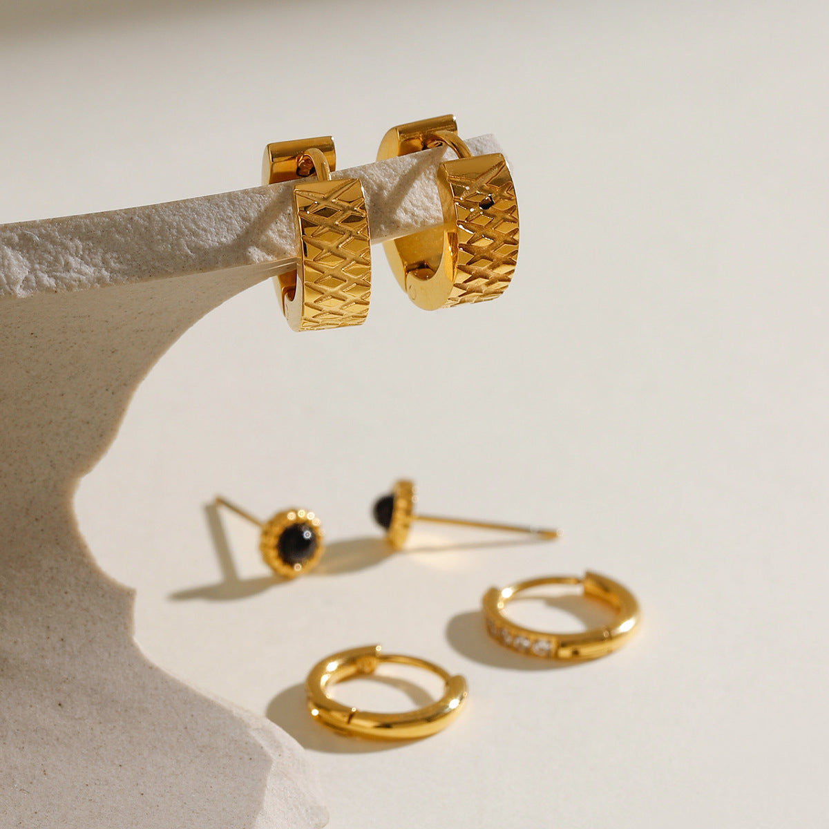 Trio of gold earrings — wide hoops, pave hoops, and gemstone studs — arranged on a glass block.