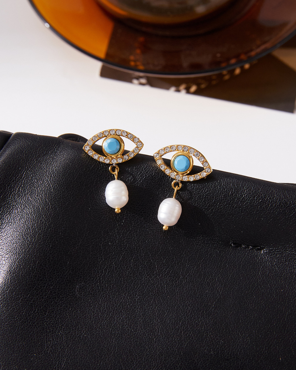Close-up of evil eye earrings with crystal outline and dangling pearl on black leather bag.