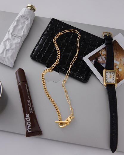 Gold chain necklace, black wallet, and watch on a light gray surface with a Rhode product.
